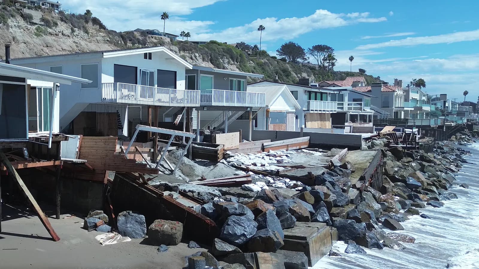 Beachfront homes at Capistrano Beach with collapsed decks and broken concrete above a rock wall, and surf reaching the base of the rocks.
